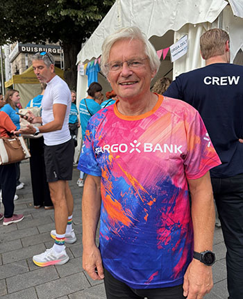 Ein älterer Mann steht lächelnd in einem bunten T-Shirt mit dem Logo der TARGO BANK. Im Hintergrund sind Menschen und ein Zelt zu sehen. Einige tragen sportliche Kleidung.