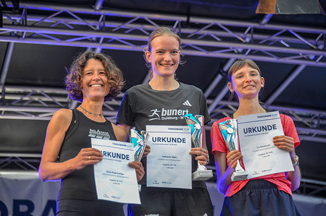 Drei Frauen stehen auf einer Bühne und halten ihre Urkunden in der Hand. Die Frau in der Mitte trägt ein schwarzes T-Shirt mit dem Schriftzug 'bunerit Duisburg'. Die anderen zwei Frauen sind links und rechts von ihr, eine trägt ein schwarzes Shirt und die andere ein rotes Shirt. Alle lächeln und scheinen stolz auf ihre Auszeichnungen zu sein.