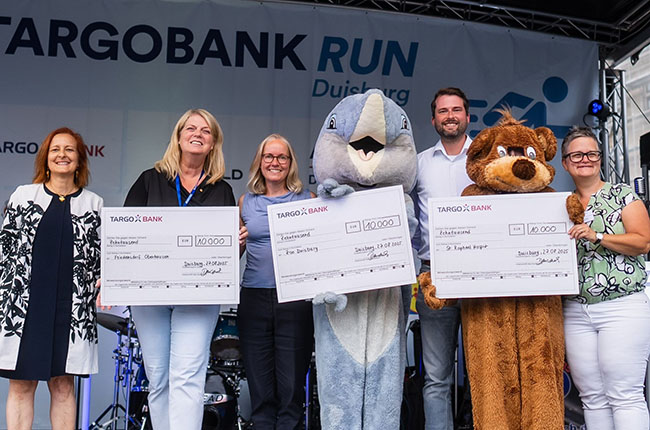 Bei der TARGOBANK RUN-Veranstaltung in Duisburg stehen fünf Personen auf einer Bühne mit großen Schecks. Drei Frauen und ein Mann halten die Schecks, während eine Person in einem Maskottchenkostüm eines Delphins und ein weiteres in einem Bärenkostüm neben ihnen stehen. Im Hintergrund sind Banner mit dem Logo der TARGOBANK und eine Band zu sehen.
