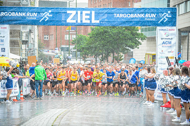 Eine große Gruppe von Läufern startet beim TARGOBANK RUN in Duisburg. Es regnet stark, während die Teilnehmer auf den Startschuss warten. Über ihnen hängt ein blaues Banner mit dem Wort 'ZIEL'. Cheerleader in blauen Outfits stehen am Rand und feuern die Läufer an.