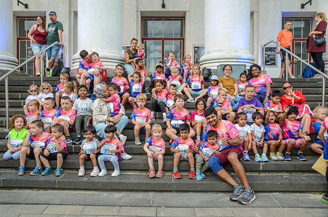 Eine Gruppe von Kindern sitzt auf den Stufen eines Gebäudes mit Säulen. Sie tragen bunte T-Shirts und Nummern auf ihren Shirts. Einige Erwachsene stehen im Hintergrund. Es sind zahlreiche Kinder verschiedenen Alters und Geschlechts zu sehen, die lachen und posieren. Die Atmosphäre wirkt fröhlich und energetisch.