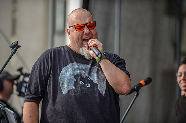 Ein Mann mit Glatze und Sonnenbrille singt ins Mikrofon. Er trägt ein schwarzes T-Shirt mit einem Grafikdesign eines Tieres vor einem Mond. Im Hintergrund sind Kameras und andere Personen zu sehen.