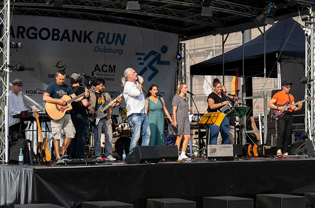 Eine Band tritt auf einer Bühne beim Argobank Run in Duisburg auf. Die Musiker spielen verschiedene Instrumente, darunter Gitarre, Schlagzeug und Saxophon. Einige Sängerinnen und Sänger stehen im Vordergrund, während im Hintergrund ein Banner mit dem Schriftzug 'Argobank Run Duisburg' zu sehen ist. Die Atmosphäre ist festlich und die Band scheint viel Energie zu haben.