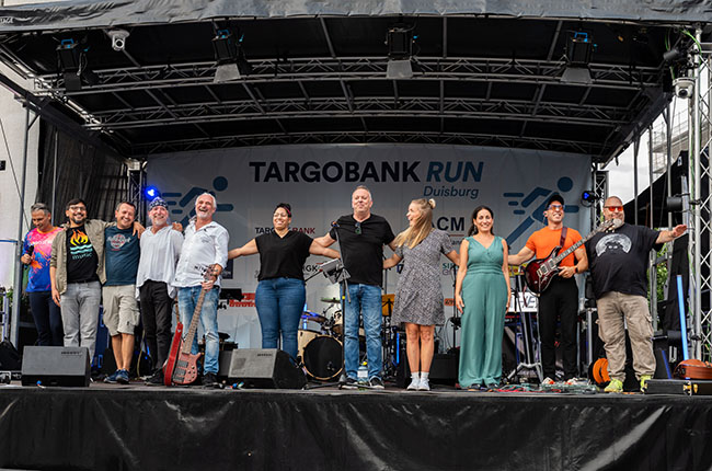 Eine Gruppe von Menschen steht auf einer Bühne und lächelt, während sie die Hände in die Luft halten. Im Hintergrund sind Musikinstrumente und eine Bühne mit einem Banner für den TARGOBANK RUN in Duisburg zu sehen. Die Gruppe umfasst Männer und Frauen in verschiedenen Outfits, einige halten Instrumente. Der gesamte Bereich ist festlich und einladend.