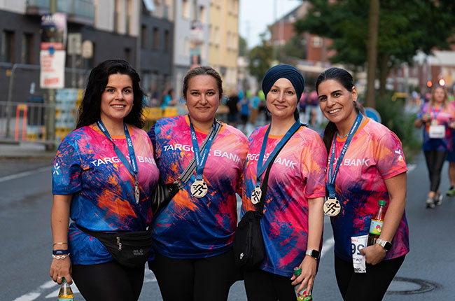 Vier Frauen stehen auf einer Straßenveranstaltung, tragen bunte T-Shirts mit dem Aufdruck 'TARGOBANK' und Medaillen um den Hals. Sie lächeln in die Kamera, während im Hintergrund andere Teilnehmer zu sehen sind.