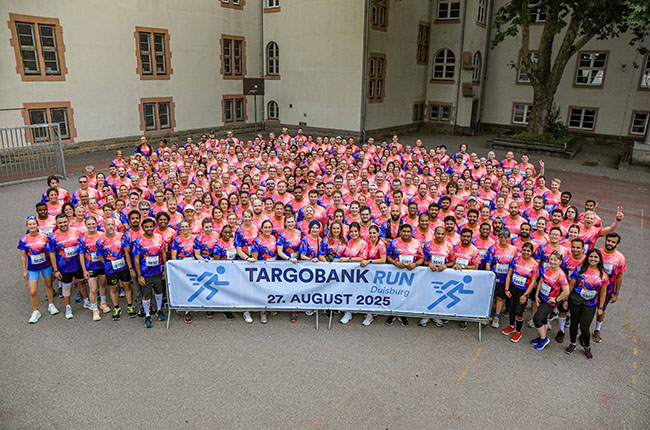 Eine große Gruppe von Menschen steht zusammen im Freien, alle tragen einheitliche T-Shirts in Pink und Blau. Sie halten ein Banner mit dem Text 'TARGOBANK RUN Duisburg 27. AUGUST 2025'. Die Szene zeigt eine fröhliche Atmosphäre und viele Personen heben ihre Hände in die Luft.