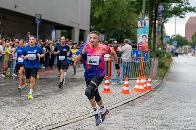 Ein Mann mit einer pink-blauen Laufbekleidung und der Startnummer 5608 sprintet auf einer nassen Straße. Im Hintergrund laufen weitere Teilnehmer, während Zuschauer an der Strecke stehen. Es gibt orangefarbene Verkehrshütchen und Werbeschilder in der Umgebung.