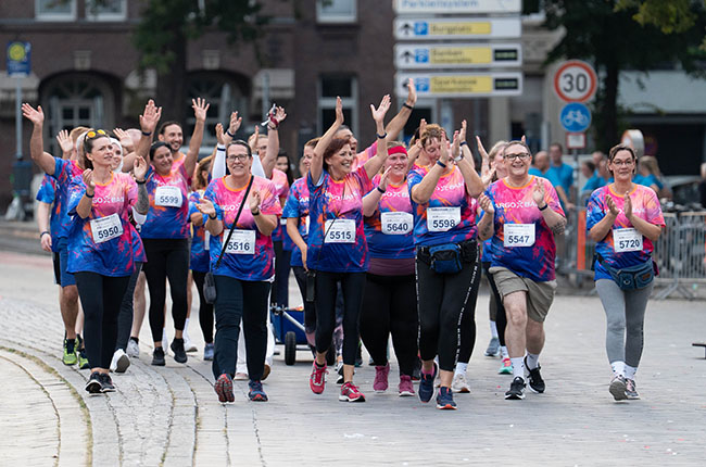 Eine Gruppe von fröhlichen Läufern in bunten T-Shirts marschiert zusammen. Sie tragen Startnummern und winken in die Kamera. Die Umgebung zeigt Straßenbeschilderungen und eine urbane Kulisse.