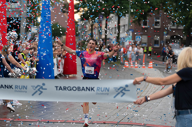 Ein männlicher Läufer mit der Startnummer 5702 durchquert die Ziellinie beim TARGOBANK RUN in Duisburg. Um ihn herum feiern Zuschauer und Konfetti fliegt durch die Luft. Im Hintergrund sind bunte Säulen und eine große Menschenmenge zu sehen.