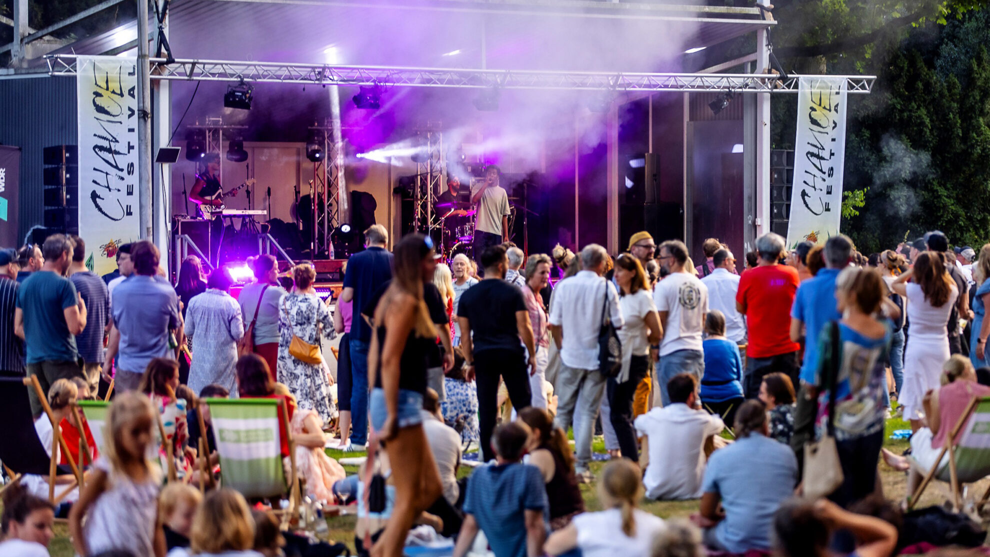 Eine Menschenmenge bei einem Musikfestival steht vor einer Bühne mit bunten Lichtern und Nebel. Ein Musiker spielt Gitarre, während ein Schlagzeuger im Hintergrund sitzt. Viele Zuschauer sitzen auf dem Boden oder stehen in Gruppen und genießen die Musik. Bunte Banner mit dem Schriftzug "CHANCE FESTIVAL" sind sichtbar. Die Atmosphäre ist lebhaft und festlich.