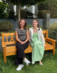 Zwei Frauen sitzen auf einer Holzbank im Freien. Die Frau links trägt ein gestreiftes Oberteil und schwarze Hosen, die Frau rechts ein grünes Kleid mit einer weißen Bluse. Im Hintergrund sind grüne Pflanzen und ein grauer Hintergrund sichtbar.