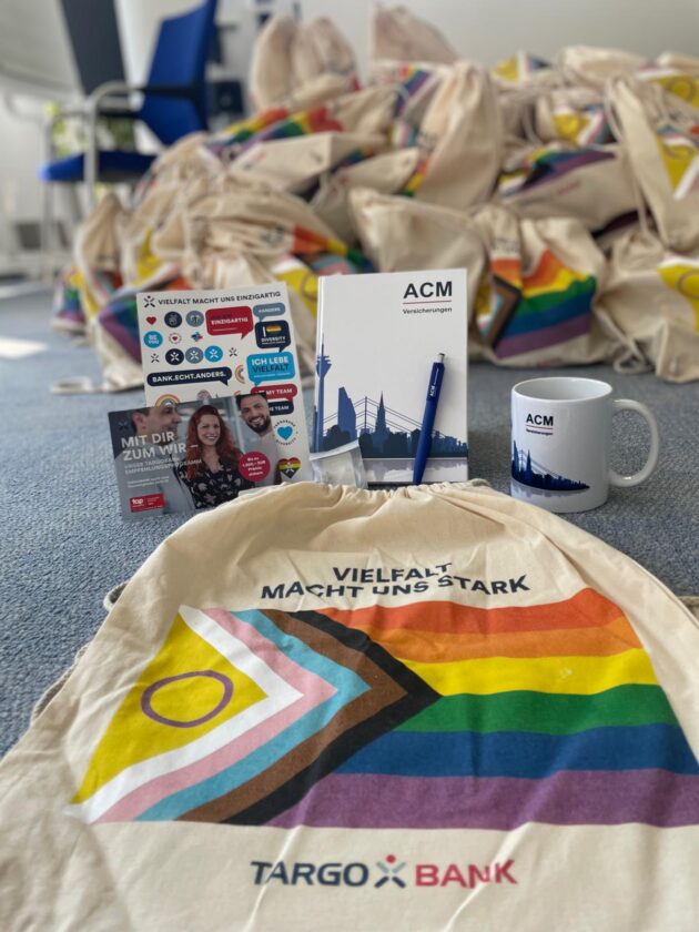 Ein Tisch mit verschiedenen Gegenständen: Eine Tragetasche mit einem Regenbogenmuster, ein Notizbuch von ACM, ein Stift, eine Tasse mit dem ACM-Logo und ein Flyer. Im Hintergrund sieht man einen großen Stapel ähnlicher Tragetaschen. Der Text 