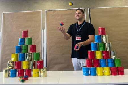 Ein Mann steht in einem Raum und jongliert mit bunten Bällen. Vor ihm befinden sich zwei pyramidale Stapel aus bunten Dosen in Rot, Grün, Blau und Gold. Der Mann trägt ein schwarzes T-Shirt und hat ein Namensschild um den Hals. Im Hintergrund sind braune Wände und einige rote Stühle sichtbar.