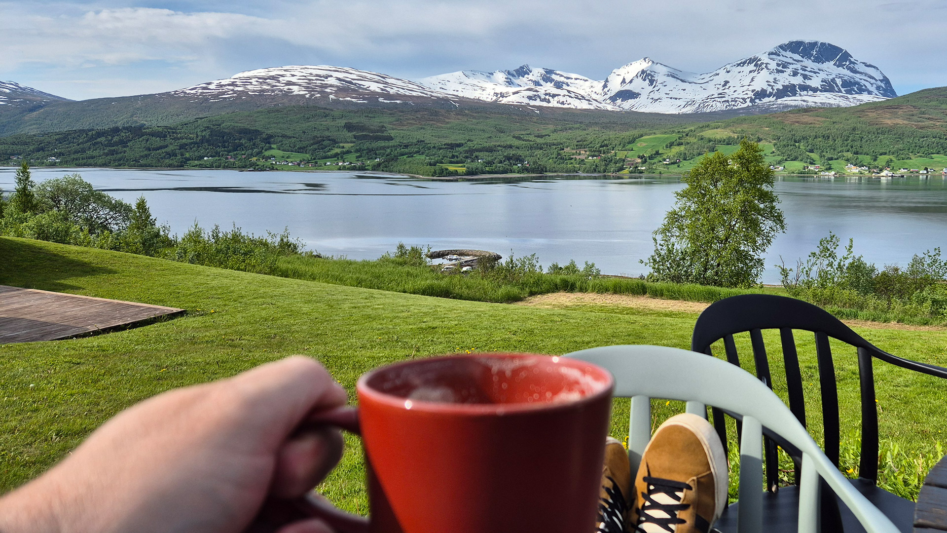 Ein Blick auf einen ruhigen See, umgeben von grünen Hügeln und schneebedeckten Bergen im Hintergrund. Im Vordergrund hält eine Person eine rote Tasse, während sie auf einem Stuhl sitzt. Ihre Füße sind auf einem anderen Stuhl platziert. Die Landschaft zeigt eine Mischung aus grasbewachsenen Flächen und Bäumen, die den See umgeben.
