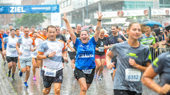 Eine große Gruppe von Läufern, die in einer Stadt bei Regen an einem Marathon teilnehmen. Einige Läufer tragen Nummern auf ihren Trikots. Die Stimmung ist fröhlich, mit einer Frau in einem blauen Shirt, die jubelnd die Arme hochhebt. Im Hintergrund ist ein Banner mit der Aufschrift 'ZIEL' zu sehen.