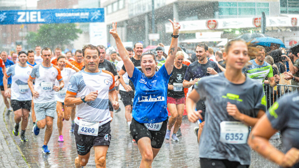 Eine große Gruppe von Läufern, die in einer Stadt bei Regen an einem Marathon teilnehmen. Einige Läufer tragen Nummern auf ihren Trikots. Die Stimmung ist fröhlich, mit einer Frau in einem blauen Shirt, die jubelnd die Arme hochhebt. Im Hintergrund ist ein Banner mit der Aufschrift 'ZIEL' zu sehen.