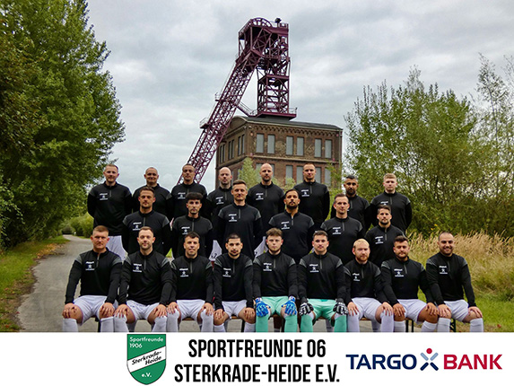 Eine Fußballmannschaft steht auf einem Weg vor einem alten Gebäude mit einem Förderturm im Hintergrund. Die Spieler tragen schwarze Trikots mit weißen Hosen. Im Vordergrund sitzen die Spieler, während die anderen in zwei Reihen dahinter stehen. Am unteren Bildrand sind die Logos von Sportfreunde 06 Sterkrade-Heide e.V. und TARGO BANK zu sehen.