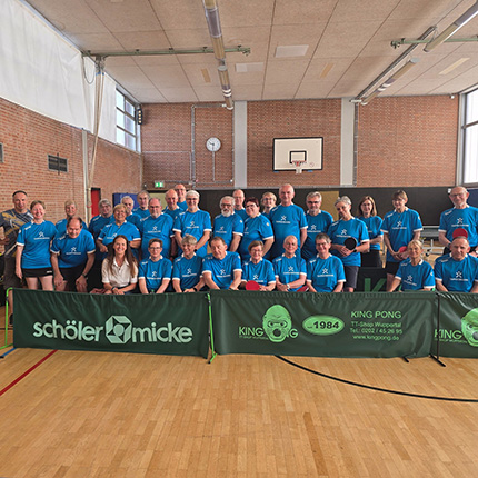 Eine große Gruppe von Menschen in blauen T-Shirts steht zusammen in einer Sporthalle. Im Hintergrund sind Basketballkörbe sichtbar, und es gibt ein grünes Tischtennisnetz mit der Aufschrift 'KING PONG'. Die Teilnehmer scheinen an einem Tischtennisturnier oder einer Veranstaltung teilzunehmen. Einige Personen lächeln, während sie posieren, und es gibt auch eine Person in der Mitte, die eine weiße Bluse trägt.