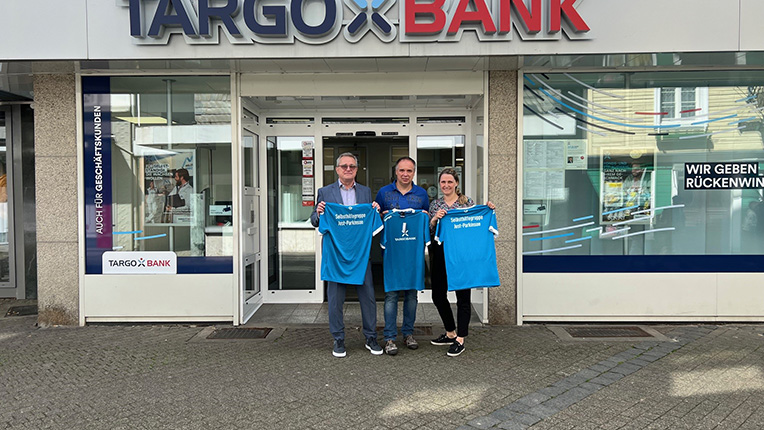 Drei Personen stehen vor der TARGOBANK-Filiale, die im Hintergrund sichtbar ist. Sie halten blaue T-Shirts mit Aufschriften in den Händen. Die Person links trägt Brille und hat graue Haare, die Person in der Mitte hat kurze Haare und ist lässig gekleidet, die Person rechts hat lange Haare und trägt ein T-Shirt. Die Eingangstüren der Bank sind offen und im Hintergrund sind Werbeflächen sichtbar.
