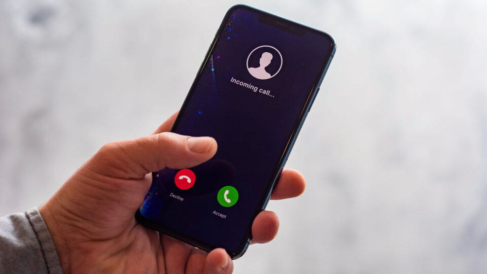 Ein Smartphone mit einem eingehenden Anruf wird in einer Hand gehalten. Der Bildschirm zeigt einen Anruf mit der Aufschrift "Eingehender Anruf..." und zwei Schaltflächen: eine rote zum Ablehnen und eine grüne zum Annehmen des Anrufs.