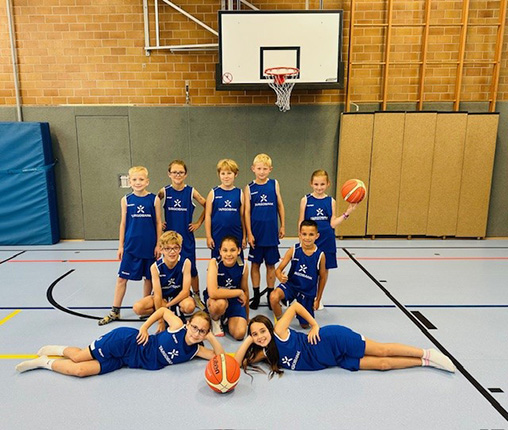 Eine Gruppe von Kindern in blauen Basketballtrikots steht und liegt auf dem Boden eines Sportplatzes. Sie halten Basketballs und posieren fröhlich vor einem Basketballkorb. Die Umgebung ist hell mit einer Ziegelwand im Hintergrund.