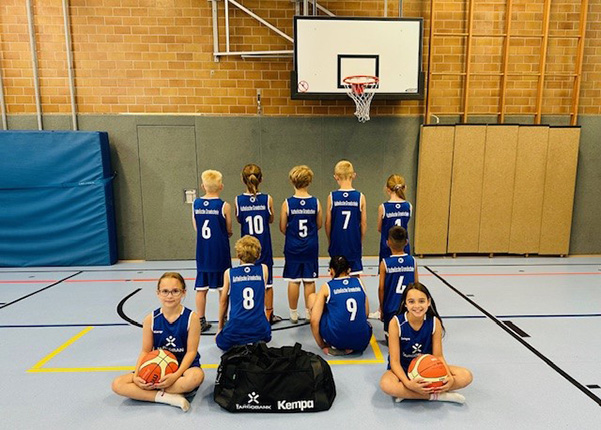 Eine Gruppe von Basketballspielern steht in blauen Trikots mit Nummern auf dem Rücken. Vor ihnen sitzen zwei Mädchen mit Basketballs, während sie eine Sporttasche mit dem Kempa-Logo haben. Im Hintergrund ist ein Basketballkorb und eine Sporthalle sichtbar.