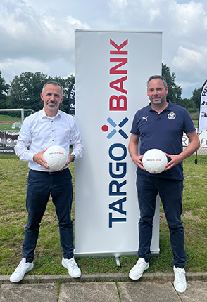 Zwei Männer stehen vor einem Banner mit dem Logo der TARGO BANK. Der Mann links trägt ein weißes Hemd und hält einen Fußball, während der Mann rechts ein sportliches Outfit trägt und ebenfalls einen Fußball hält. Im Hintergrund sind Fußballtore und Bäume sichtbar.