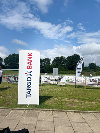 Ein Banner der TARGO BANK steht auf einem Rasenfeld, umgeben von weiteren Werbebannern. Im Hintergrund sind grüne Bäume und ein blauer Himmel mit einigen Wolken zu sehen.