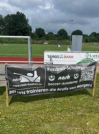 Ein Werbebanner für die nash Fussball-Schule steht auf einem Sportplatz. Es zeigt den Text 'nash Soccer-Academy' und einen QR-Code. Im Hintergrund sieht man ein Fußballfeld mit Toren und einigen Markierungen. Es gibt auch eine TARGOBANK-Werbung und eine grüne Fläche im Bild.