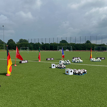 Ein Fußballfeld mit verschiedenen Nationalflaggen (Deutschland, Türkei, Frankreich) und mehreren Fußballkästen. Auf dem Feld liegen viele Fußbälle verteilt. Der Himmel ist bewölkt.