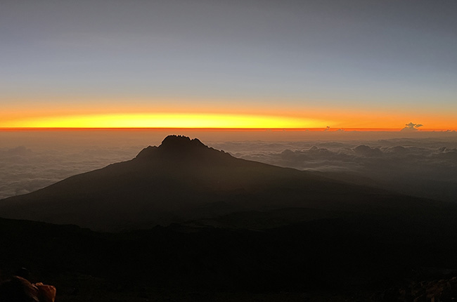 Ein Sonnenuntergang über einer Berglandschaft. Der Gipfel eines Berges ist silhouettiert gegen den orangefarbenen Himmel, der sich über einer Wolkendecke erstreckt. Die Farben des Himmels ändern sich von Blau zu Orange und Gelb, während die Wolken sanft unter dem Berg liegen.