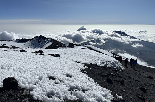 Eine schneebedeckte Berglandschaft mit Wolken, die über den Gipfel ziehen. Wanderer sind auf einem schmalen Pfad zu sehen, während der Himmel klar und blau ist.