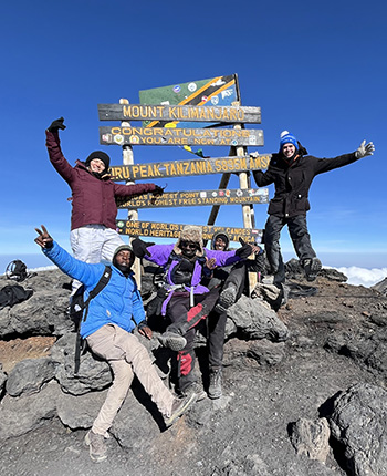 Eine Gruppe von fünf Menschen steht auf dem Gipfel des Mount Kilimanjaro. Sie posieren fröhlich vor einem Schild, das 