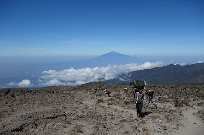Eine Wandergruppe auf einem schroffen Weg in einer bergigen Landschaft. Im Hintergrund erhebt sich ein großer Berg, umgeben von Wolken. Die Gruppe besteht aus mehreren Personen, die Rucksäcke tragen, während sie den steinigen Pfad entlanggehen. Die Atmosphäre ist klar und sonnig, mit einem blauen Himmel und wenigen Wolken.