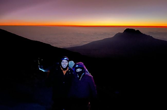 Ein Paar steht in warmer Kleidung vor einem farbenfrohen Sonnenaufgang über einer Berglandschaft. Im Hintergrund ist ein Berg sichtbar, während Wolken darunter liegen.