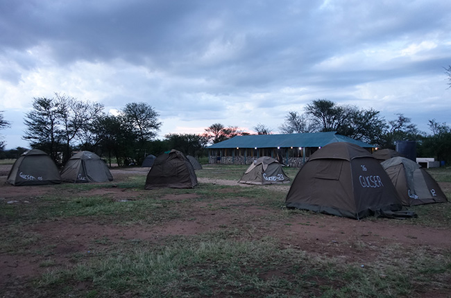 Eine Campingplatzszene mit mehreren braunen Zelten auf einem erdigen Boden. Im Hintergrund ist ein Gebäude mit einem grünen Dach und Bäumen zu sehen. Der Himmel ist bewölkt, und es gibt Anzeichen von Sonnenuntergang.