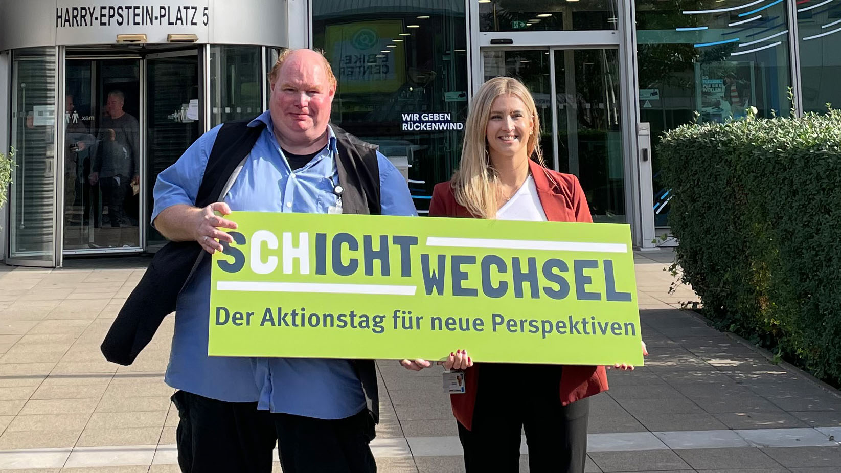 Zwei Personen stehen vor einem Gebäude mit dem Schild 'SCHICHTWECHSEL - Der Aktionstag für neue Perspektiven'.