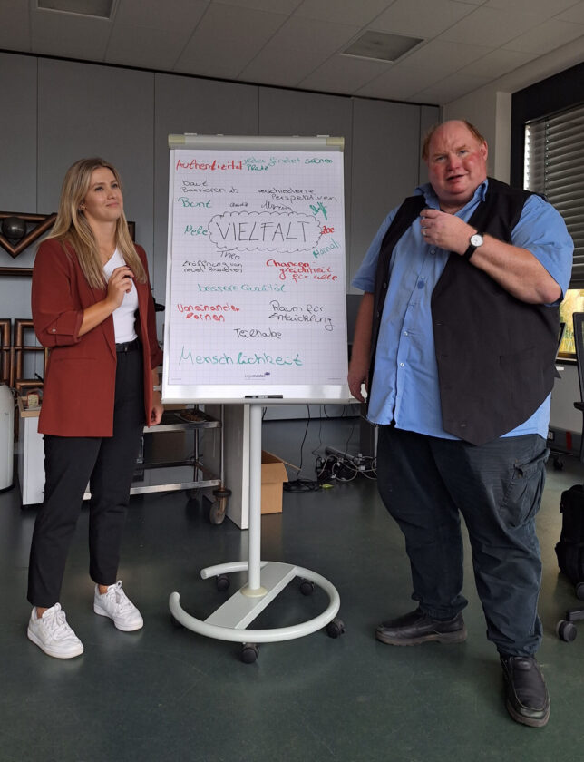 Eine Frau mit blonden Haaren in einem roten Blazer und ein Mann mit kurz geschnittenen Haaren in einem blauen Hemd und schwarzer Weste stehen neben einem Flipchart. Auf dem Flipchart steht 