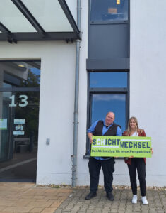 Zwei Personen stehen vor einem Gebäude mit der Nummer 13. Sie halten ein großes Schild mit der Aufschrift "SCHICHTWECHSEL - Der Aktionstag für neue Perspektiven". Die Person links trägt eine Weste und ein blaues Hemd, während die Person rechts einen roten Blazer trägt. Im Hintergrund sind Fenster und ein Teil der Fassade des Gebäudes sichtbar.