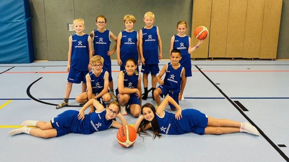 Eine Gruppe von zehn Kindern in blauen Basketballtrikots steht und posiert auf einem Basketballfeld. Einige Kinder knien, während andere stehen. Ein Mädchen liegt auf dem Boden und hält einen Basketball. Die Umgebung aussieht nach einer Sporthalle mit einem glatten Boden und Hintergrundelementen aus Schaumstoff.
