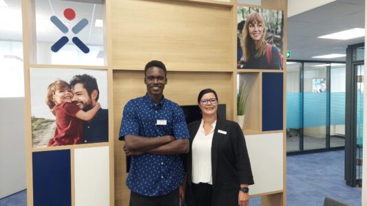 Ein Mann und eine Frau stehen in einem modernen Büro vor einer Holzwand mit Fotos. Der Mann trägt ein blaues Hemd mit Mustern und lächelt, während die Frau in einem schwarzen Blazer mit einer weißen Bluse steht. Hinter ihnen sind Bilder von einem Vater mit seiner Tochter und einer lächelnden Frau zu sehen. Das Büro hat große Fenster und eine helle Einrichtung.