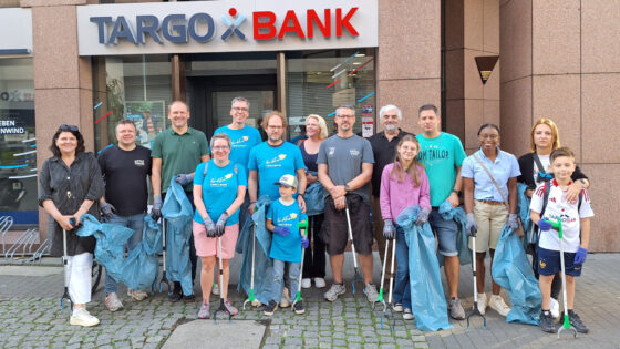 Eine Gruppe von 15 Personen steht vor der TARGOBANK in Arbeitskleidung. Die meisten tragen blaue T-Shirts und Gummihandschuhe. Einige halten Müllgreifer und Müllsäcke. Die Gruppe sieht engagiert und freundlich aus.