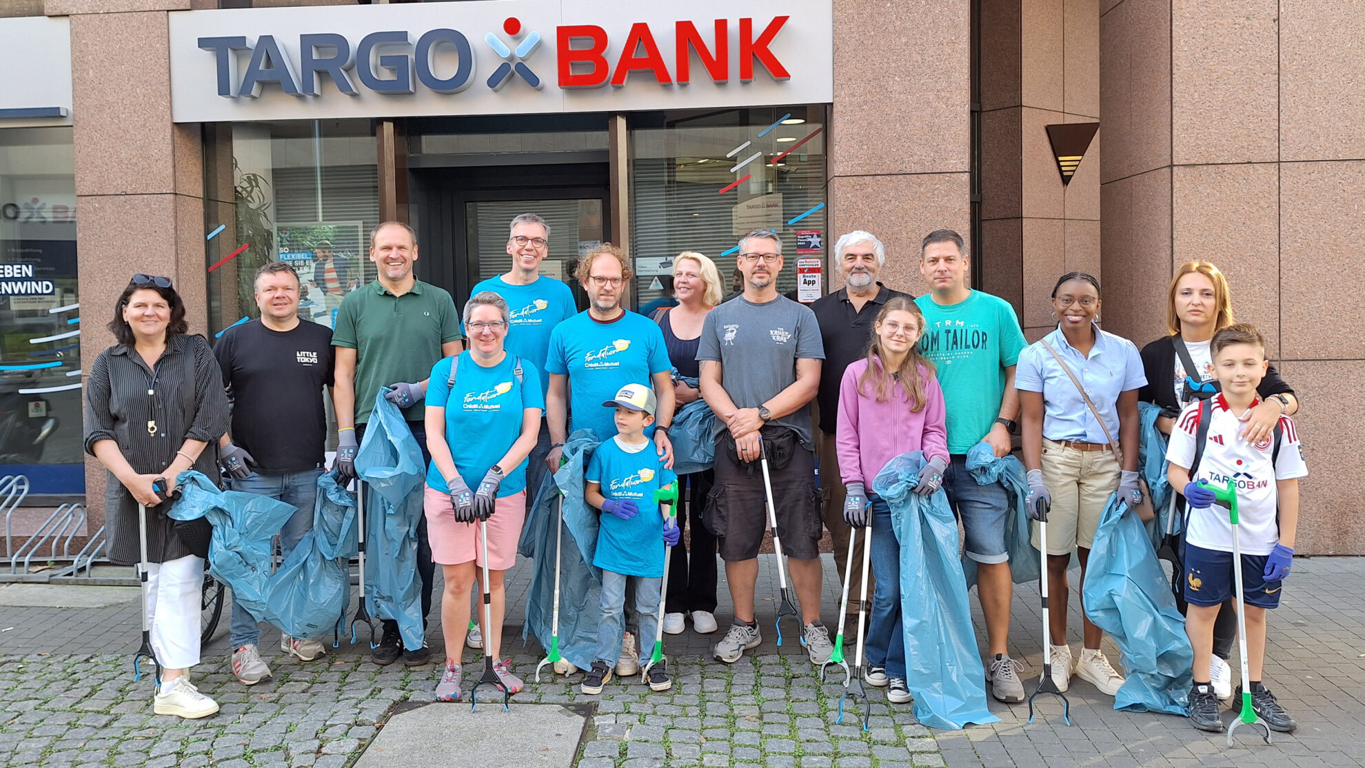 Eine Gruppe von 15 Personen steht vor der TARGOBANK in Arbeitskleidung. Die meisten tragen blaue T-Shirts und Gummihandschuhe. Einige halten Müllgreifer und Müllsäcke. Die Gruppe sieht engagiert und freundlich aus.