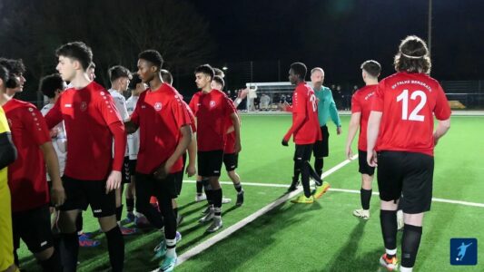 Eine Gruppe junger Fußballspieler in roten Trikots schüttelt sich die Hände nach einem Spiel. Im Hintergrund sind einige Zuschauer zu sehen, und die Szene spielt sich auf einem Kunstrasenplatz bei Nacht ab. Die Spieler tragen schwarze Shorts und es gibt einen Torwart in einem grünen Trikot. Die Nummer 12 ist auf einem der Trikots sichtbar. Die Atmosphäre ist sportlich und konzentriert.