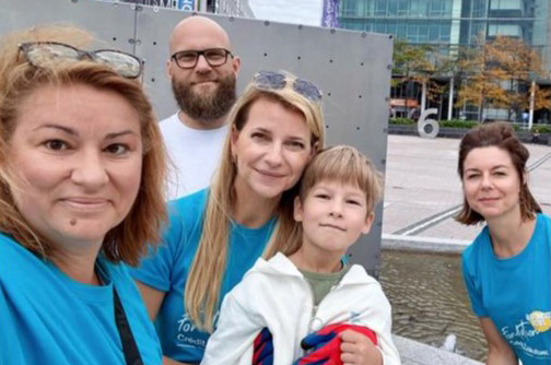 Eine Gruppe von fünf Personen posiert für ein Selfie im Freien. Alle tragen blaue T-Shirts, zwei Frauen haben lange Haare, während ein Junge in der Mitte einen weißen Hoodie trägt. Im Hintergrund ist ein Wasserbecken sichtbar.