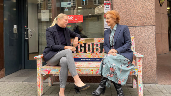 Zwei Damen sitzen auf einer Holzbank mit der Aufschrift TARGOBANK vor einer TARGOBANK Filiale.