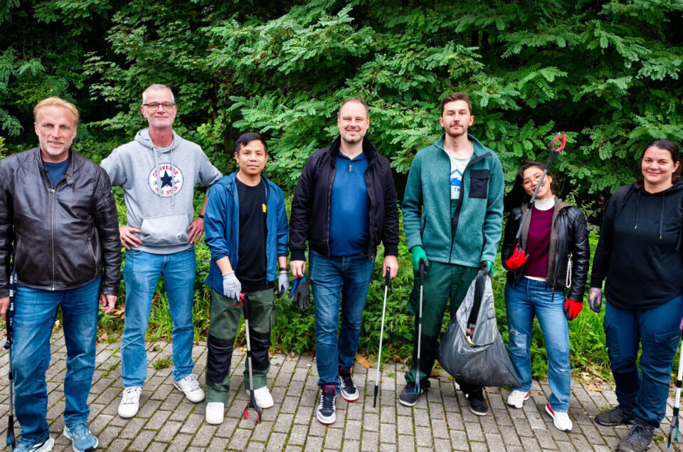 Eine Gruppe von sieben Personen steht auf einem gepflasterten Weg vor einer grünen Hecke. Sie tragen Arbeitskleidung und halten verschiedene Werkzeuge, darunter Greifzangen und einen Müllsack. Jeder hat einen freundlichen Gesichtsausdruck.