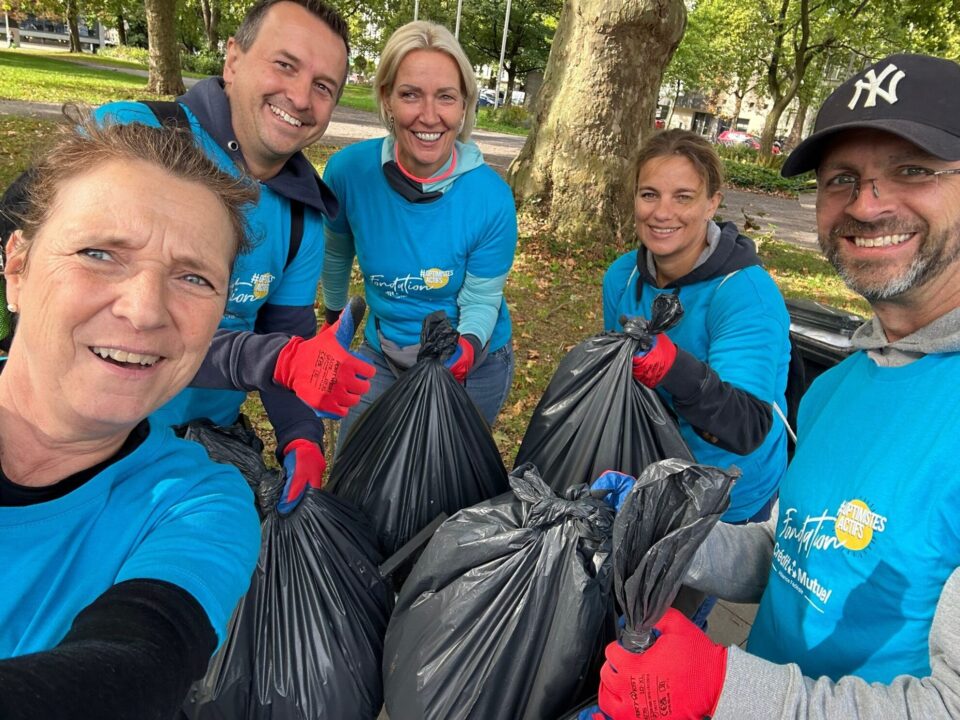 Eine Gruppe von fünf Personen in blauen T-Shirts steht im Park und hält schwarze Müllsäcke. Alle lächeln und posieren für ein Selfie. Im Hintergrund sind Bäume und ein Gehweg zu sehen.