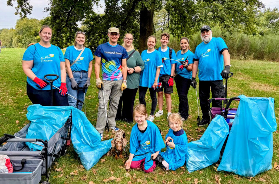 Eine Gruppe von Personen steht auf einer Wiese. Sie tragen blaue T-Shirts und arbeiten gemeinsam an einer Aufräumaktion. Im Vordergrund sind zwei kleine Mädchen in blauen T-Shirts, daneben ein Hund. Im Hintergrund sind mehrere Erwachsene, die mit Müllgreifern und Handschuhen ausgerüstet sind. Es gibt blaue Müllsäcke und Wagen, die für die Aufräumaktion verwendet werden.