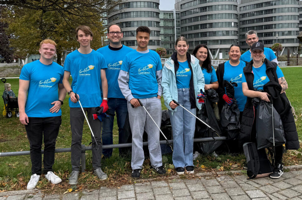 Eine Gruppe von neun Personen steht im Freien und hält Müllgreifwerkzeuge. Sie tragen blaue T-Shirts mit dem Logo der Fondation Crédit et Mutuel. Im Hintergrund sind moderne Gebäude und ein kleiner Park sichtbar. Eine Person im Hintergrund schiebt einen Kinderwagen. Die Gruppe wirkt engagiert und bereit, die Umgebung sauber zu halten.